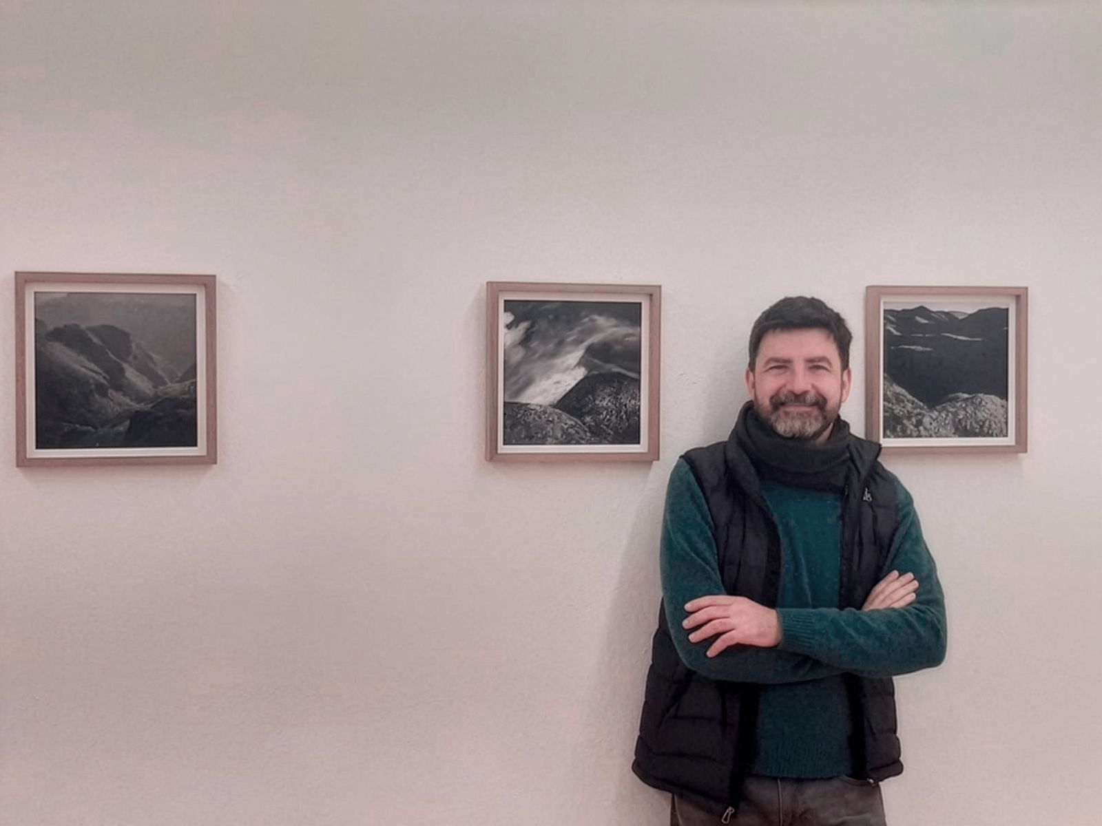 Un hombre de pie frente a tres fotografías enmarcadas de paisajes, sonriendo con los brazos cruzados.