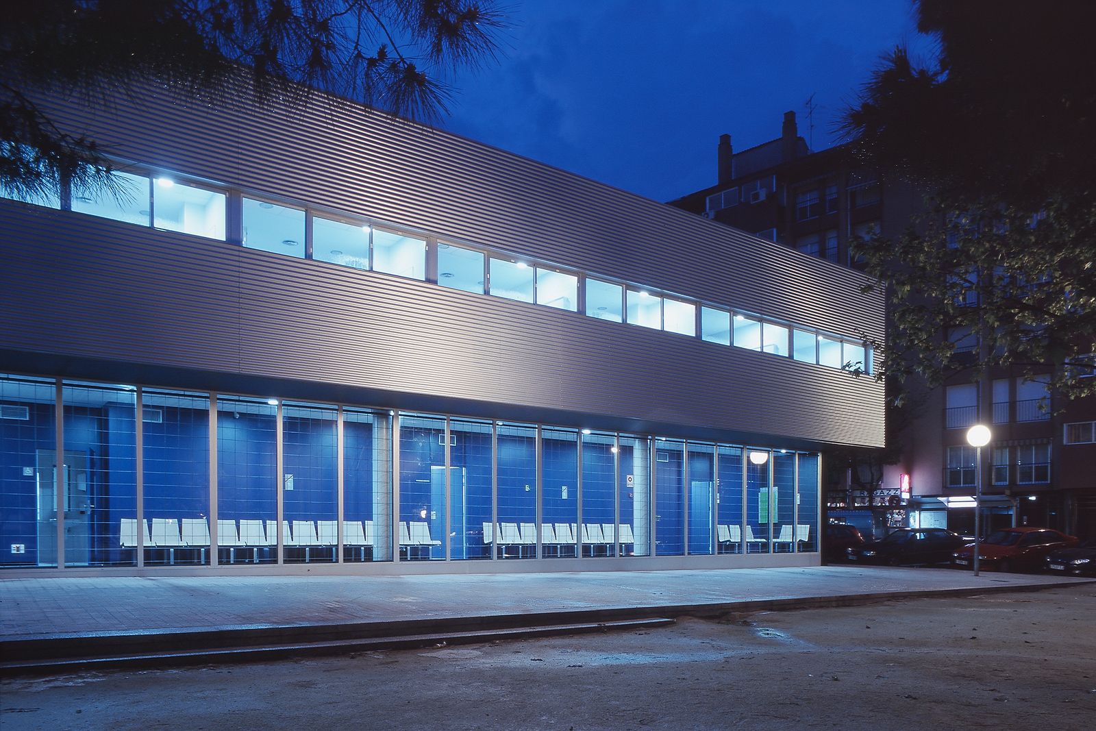 Architectural photograph at night of a modern healthcare or public center. The ground floor consists entirely of large glass windows revealing a waiting area with rows of white chairs against blue-tiled interior walls. The cantilevered upper floor is clad in horizontal corrugated metal siding with a continuous ribbon of brightly illuminated windows, set against a dark blue twilight sky and framed by nearby trees.