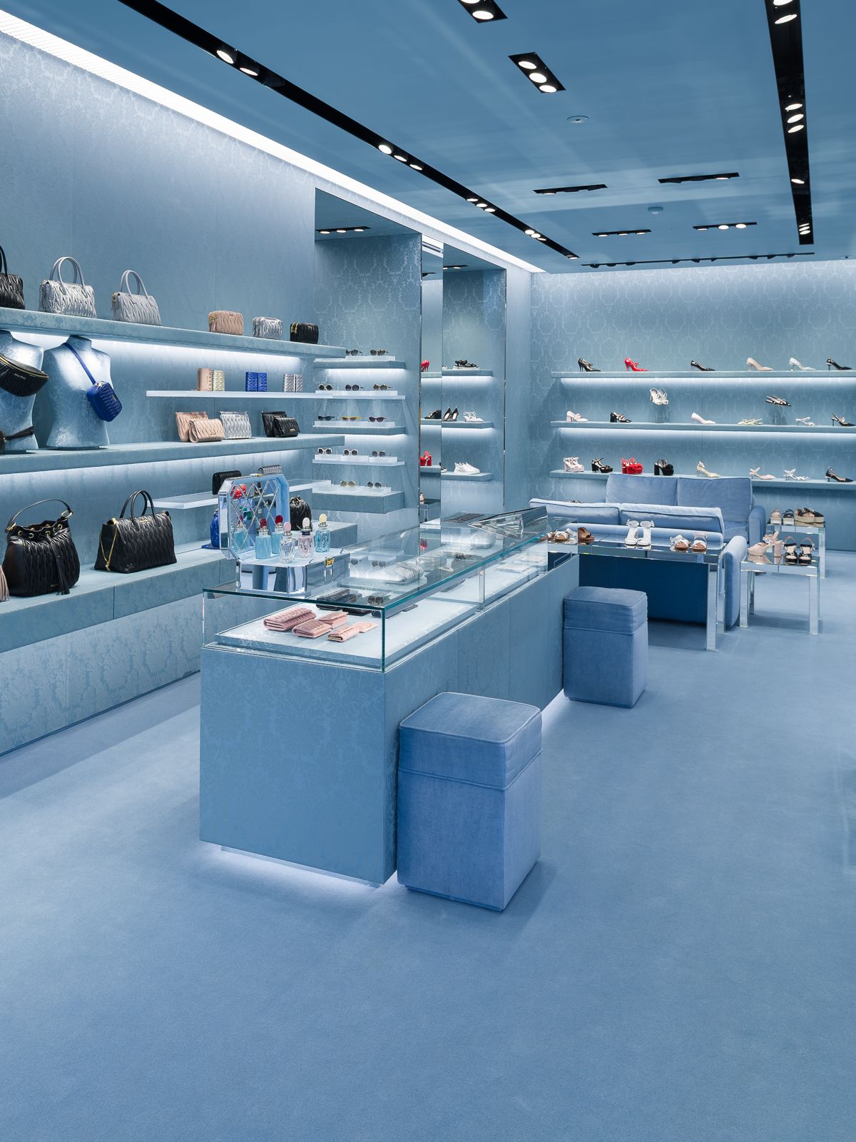Un interior elegant de botiga Miu Miu, tot en blau, mostrant prestatges d'exposició elegants amb bosses i sabates, i un taulell d'exposició de vidre amb accessoris.