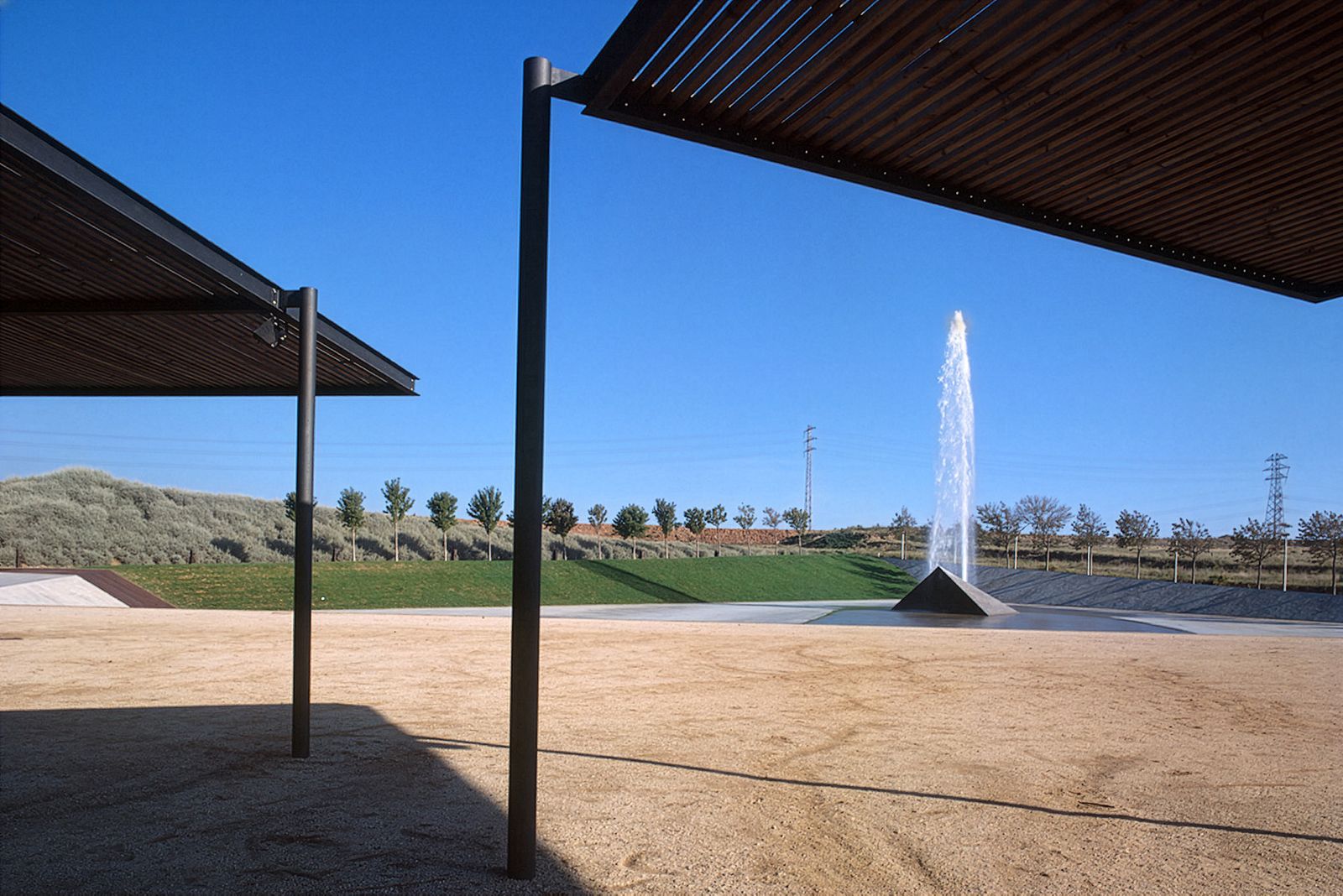 Outdoor space with two steel and wood modern shade structures and a large central jet water fountain against a clear blue sky.