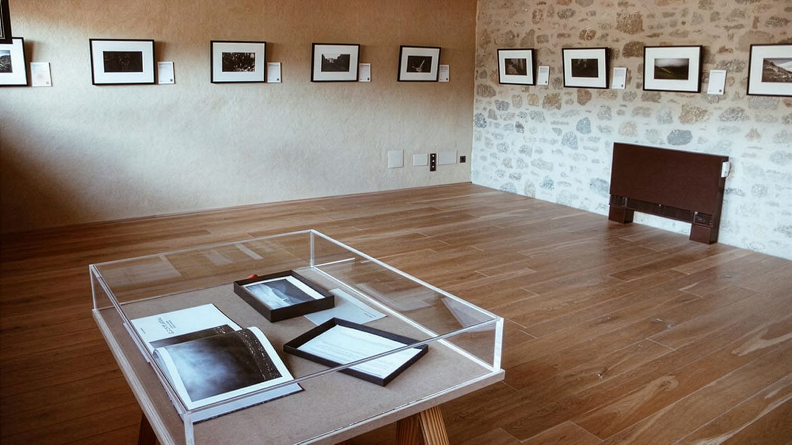 Galería con fotos enmarcadas en paredes de piedra y una mesa de exposición en el centro con fotografías y documentos.
