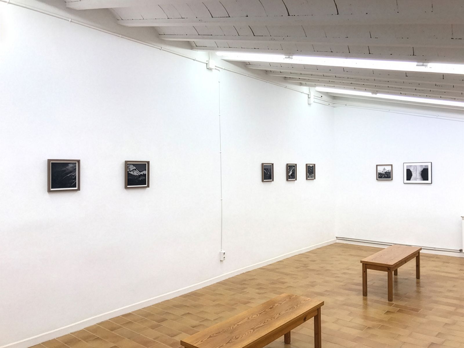 Un espacio de galería minimalista con fotografías en blanco y negro expuestas en una pared blanca y bancos de madera para sentarse.