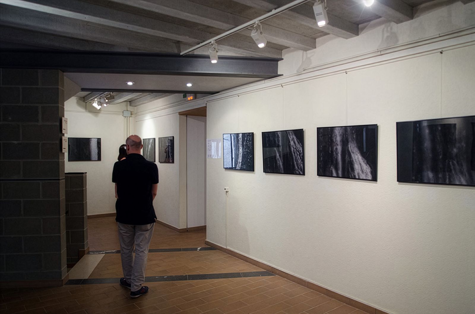 Visitante viendo fotografías enmarcadas en una galería con suelo de baldosas y vigas del techo expuestas.