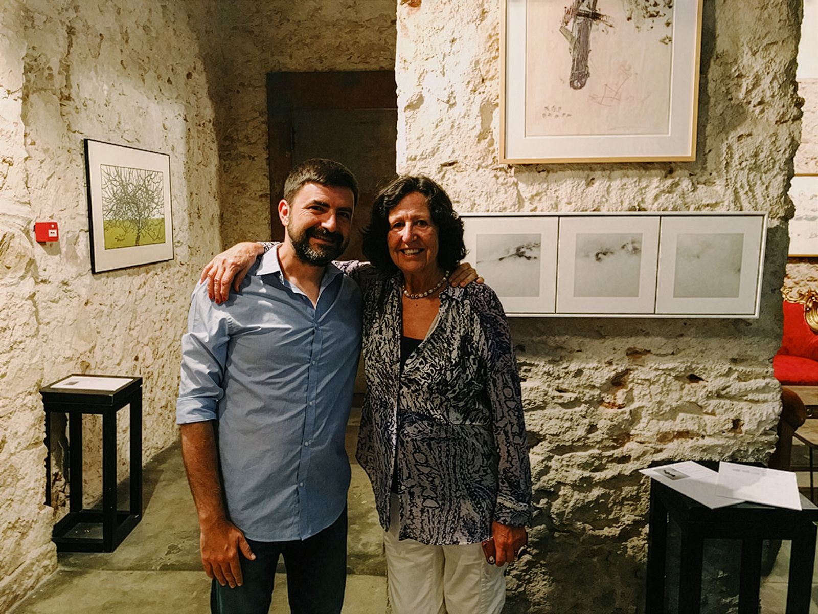 Dues persones dempeus i somrient en una galeria d'art amb obres d'art emmarcades en una paret de pedra al fons.