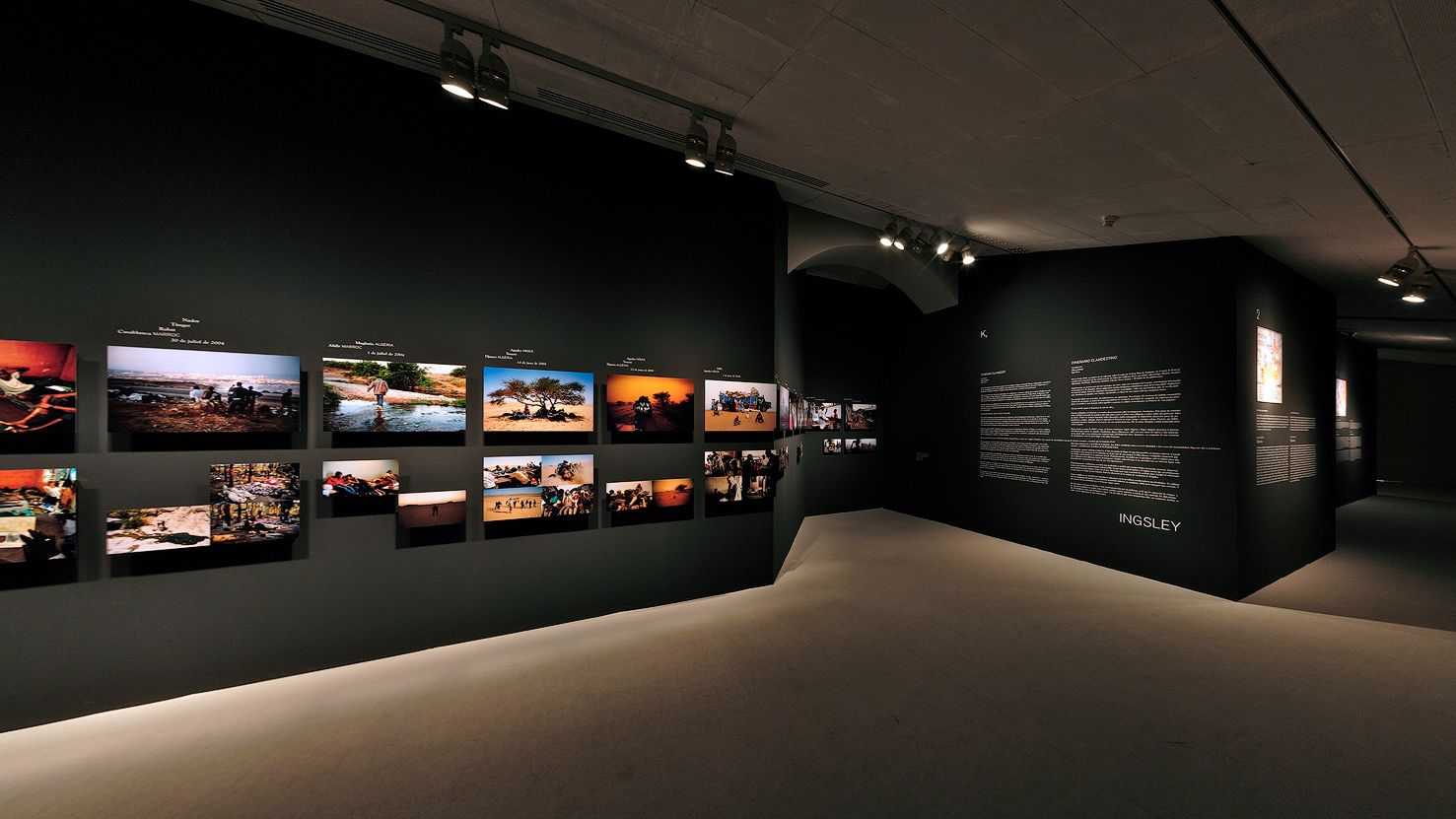Una galeria que mostra una sèrie de fotografies i panells de text descriptiu sobre parets fosques, il·luminades per focus dirigits.