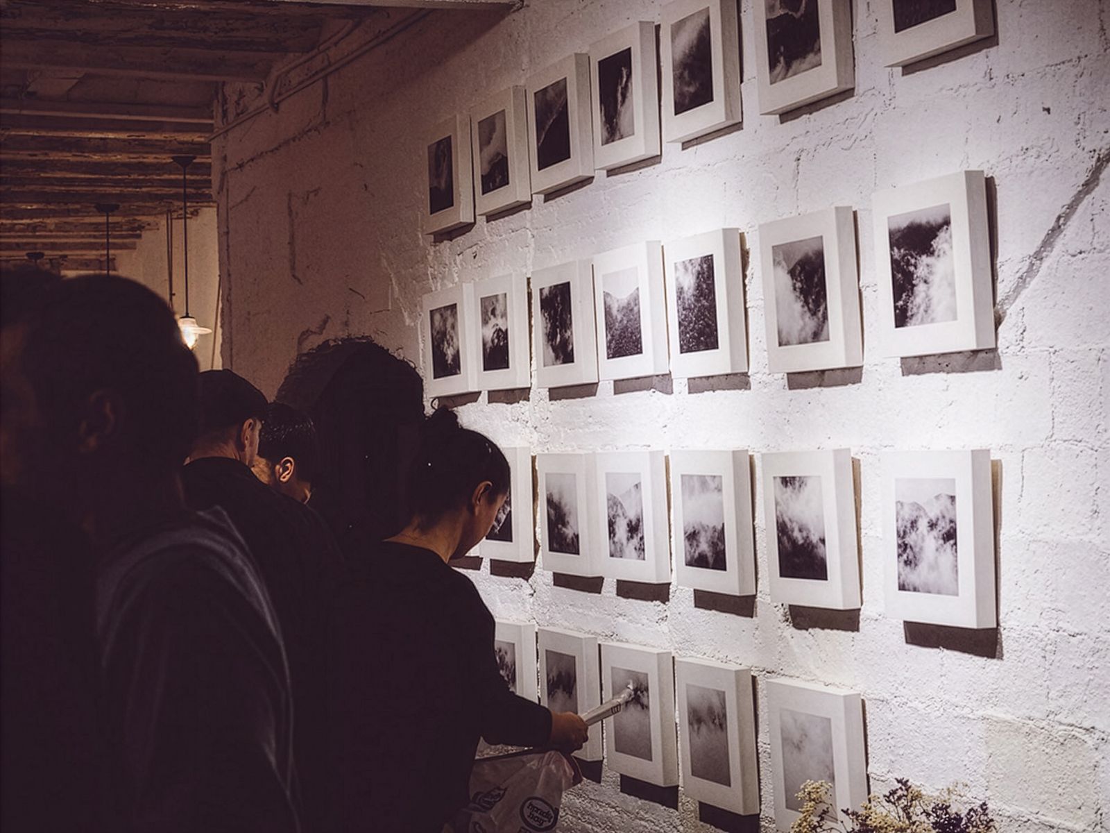 Persones veient una sèrie de fotografies en blanc i negre de núvols i boira disposades en graella en una paret de totxo blanc.