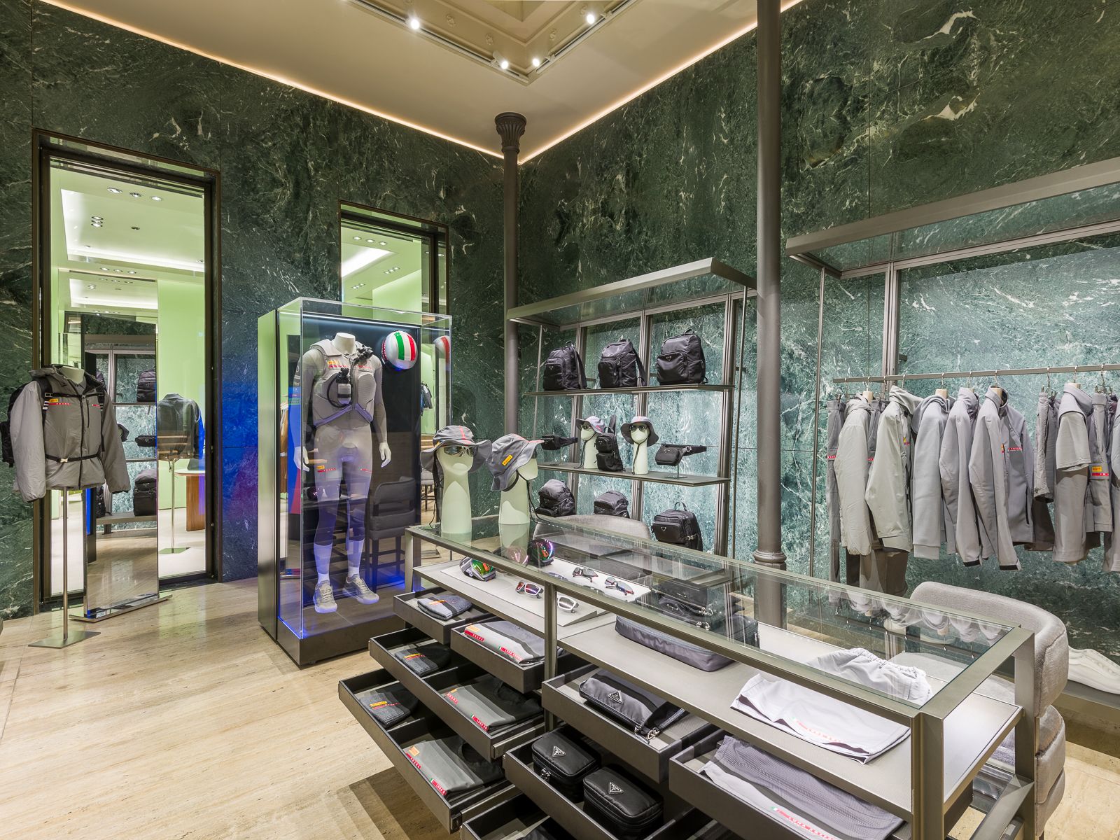 Boutique de luxe Prada amb parets de marbre verd, mostrant sabatilles esportives de dissenyador, bosses negres i accessoris en prestatges de metall elegant. Una taula de vidre mostra ulleres i barrets, mentre que un maniquí amb un vestit de rendiment està tancat en una vitrina de vidre.