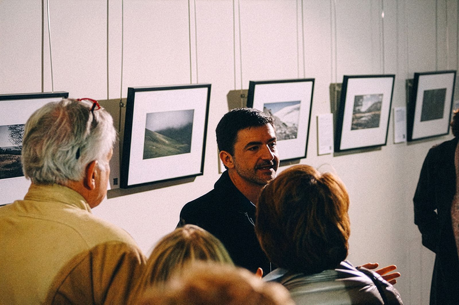 Un hombre hablando a un grupo de personas frente a una pared que muestra fotografías enmarcadas.