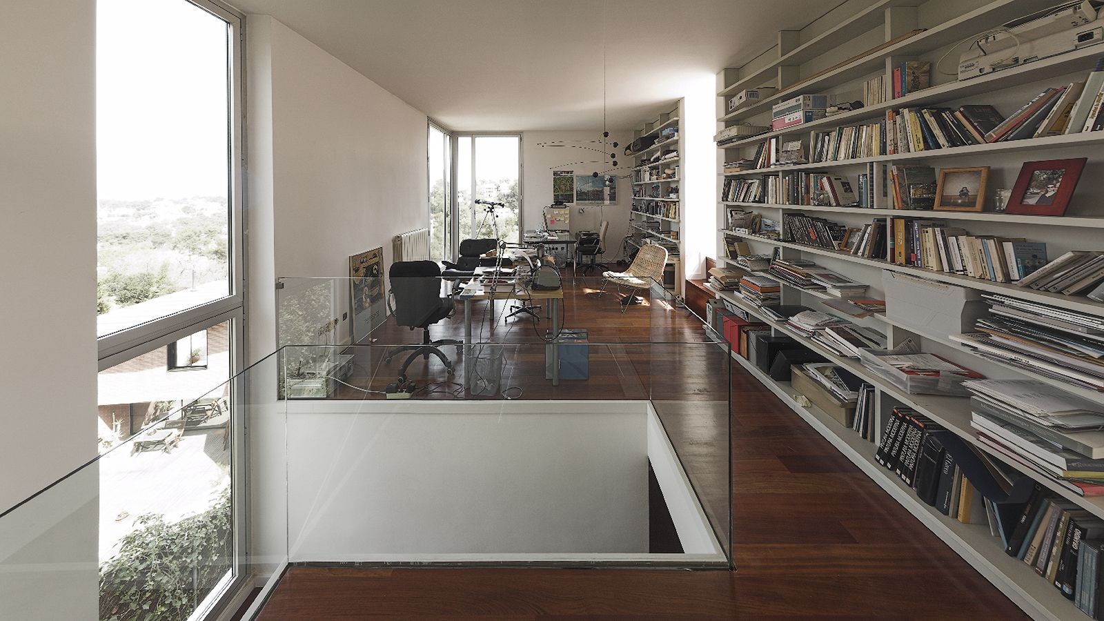 Modern home office with large windows, extensive bookshelves, and wooden floors, overlooking a green landscape.