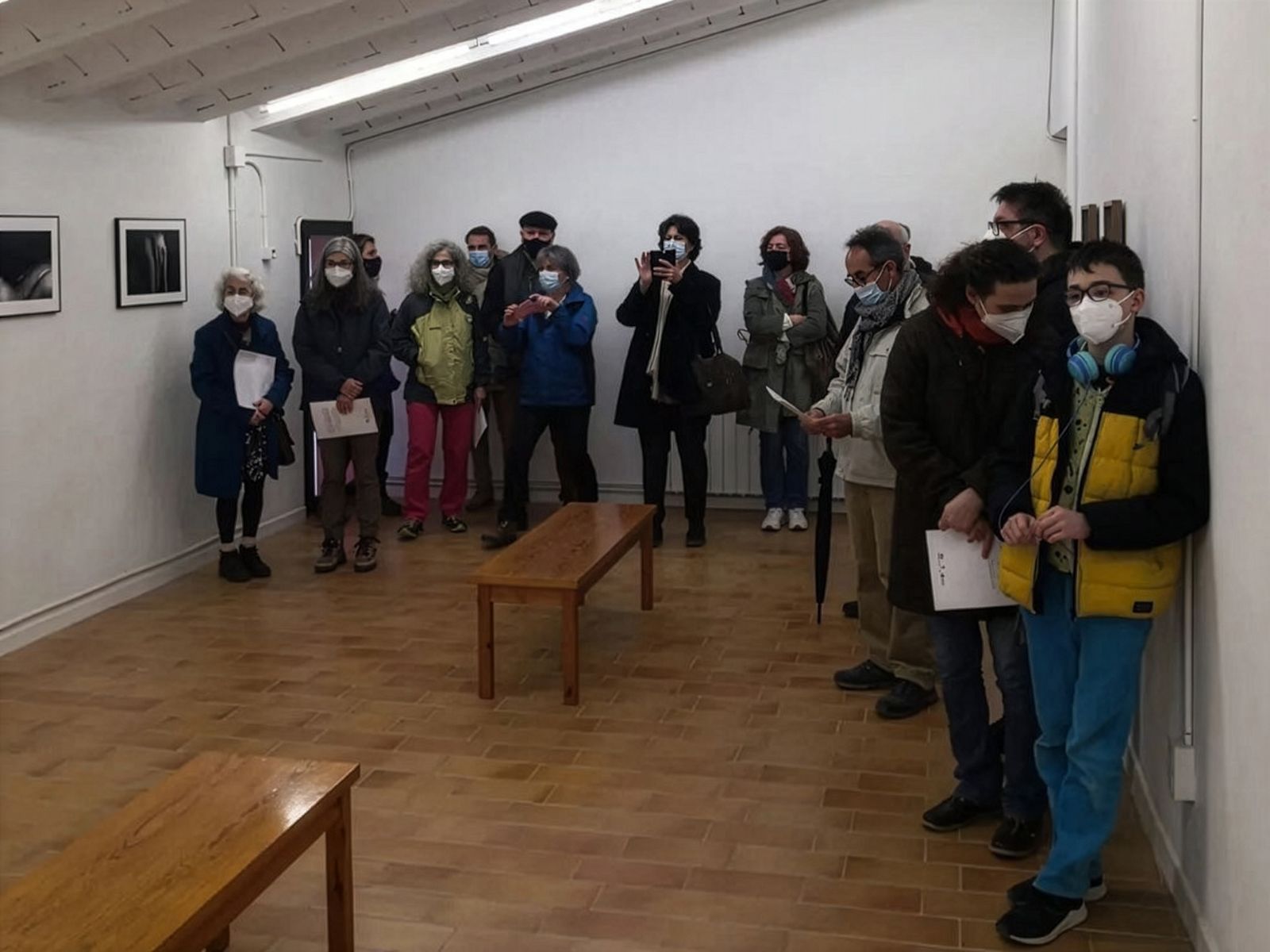 Visitantes en una galería de arte usando mascarillas quirúrgicas, algunos sosteniendo papeles y tomando fotos, observando fotografías enmarcadas en las paredes.