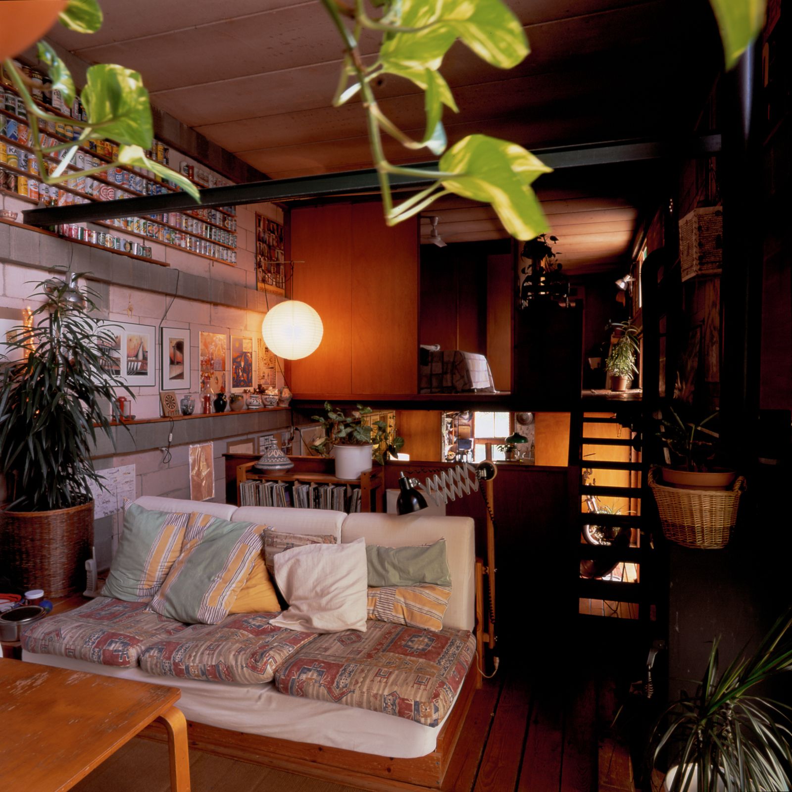 Sala d'estar acollidora amb un sofà colorit, mobles de fusta, plantes d'interior i un llum penjant blanc, amb una escala i una paret decorada amb art i objectes de col·lecció.