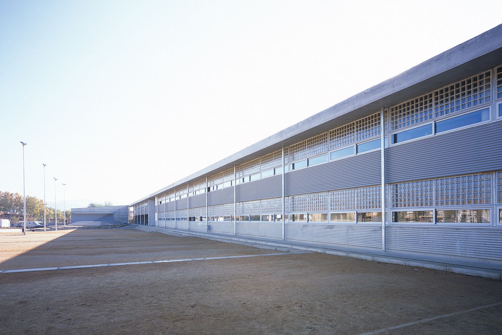 Architectural photograph of a long, linear two-story educational building. The industrial facade features alternating horizontal bands of corrugated metal cladding, continuous windows, and grids of translucent glass blocks. Slender steel columns support the clean line of a concrete roof overhang. In the foreground, a large, flat sandy schoolyard stretches out under a bright, clear blue sky.