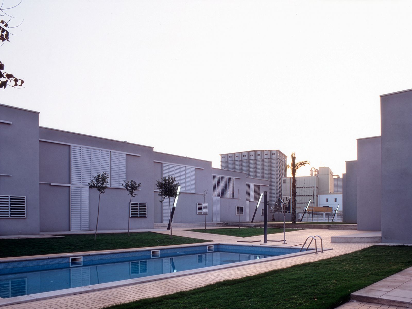 Modern residential complex with flat-roofed buildings, louvered windows, a central swimming pool, and landscaped grassy areas.