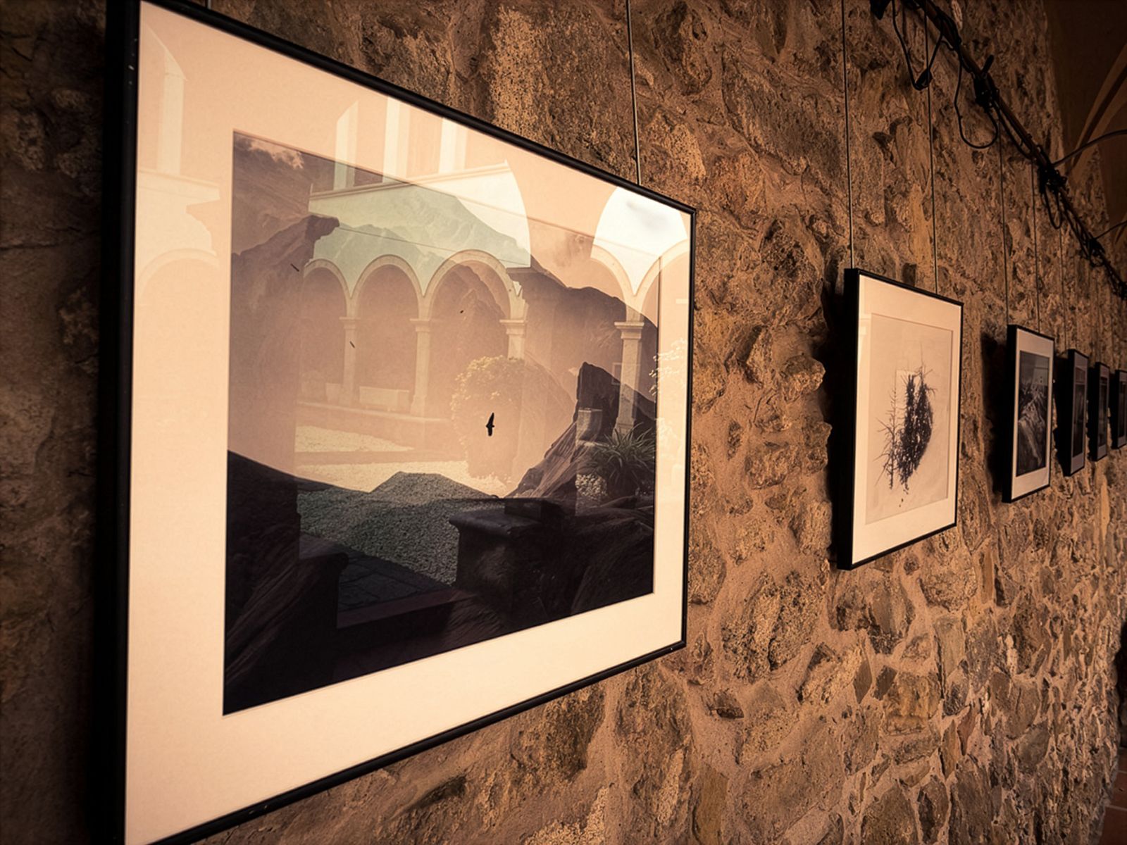Un primer pla de fotografies emmarcades penjades en una paret de pedra d'una galeria, mostrant diverses obres d'art en filera, els arcs del claustre es reflecteixen en el vidre de les fotografies.