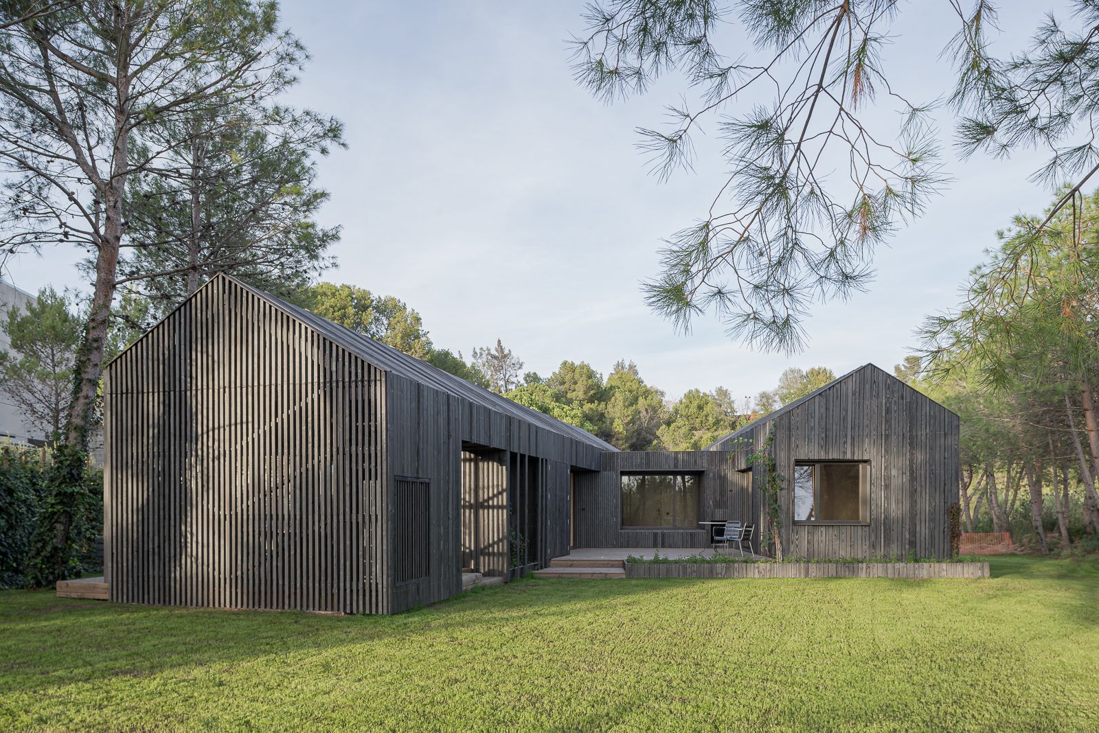 Exterior view of a modern house with two bodies, a pitched roof, dark wood cladding, large windows, and a spacious lawn surrounded by pine trees.