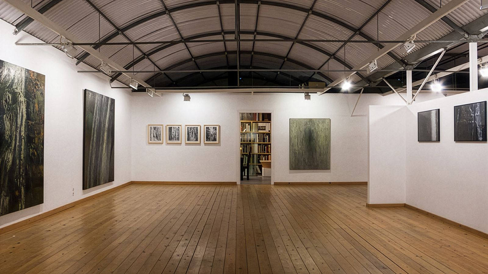 Espai d'exposició amb terra de fusta, obres d'art grans a les parets i una porta que condueix a una sala amb prestatgeries.