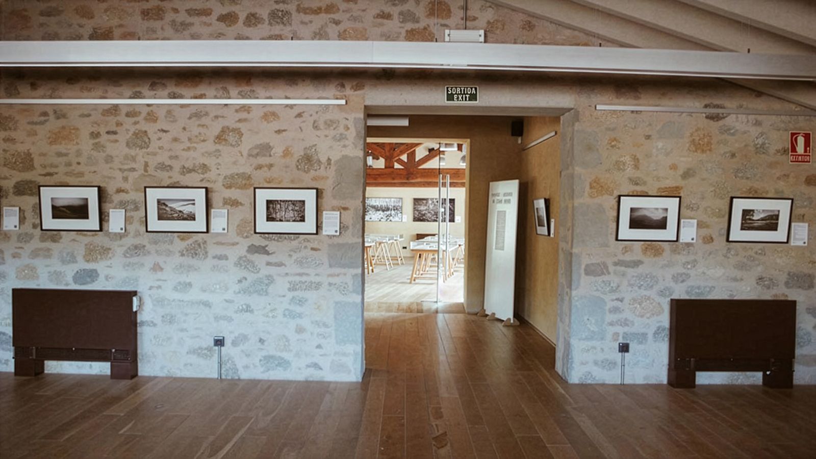 Sala con fotos enmarcadas en paredes de piedra y una entrada abierta que conduce a otra sala con más fotos enmarcadas.