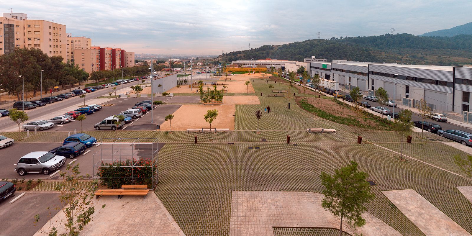 An elevated architectural landscape photograph of Foneria Park. The image displays a long, linear public plaza utilizing permeable lattice concrete grass pavers interspersed with sandy recreational zones, trees, and benches. A parking lot lines the left side next to apartment buildings, while industrial warehouses and a green hillside border the right side under a cloudy sky.