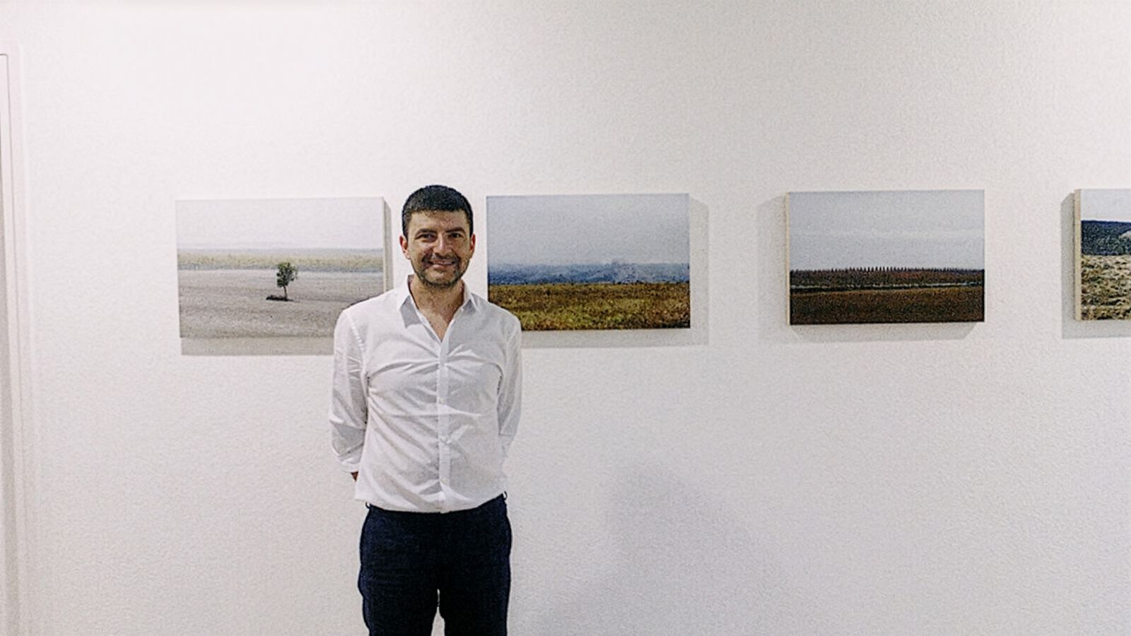 Un hombre con camisa blanca de pie frente a fotografías de paisajes en una pared de galería. Las fotografías representan varias escenas naturales, incluyendo campos y horizontes distantes.