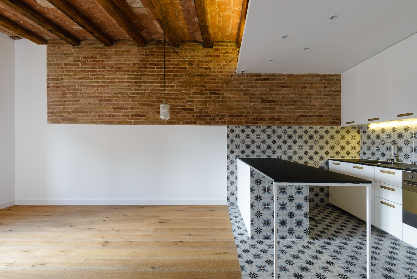 Modern open kitchen with a black work table, patterned tiles, exposed brick wall, wooden beams, and white cabinets.