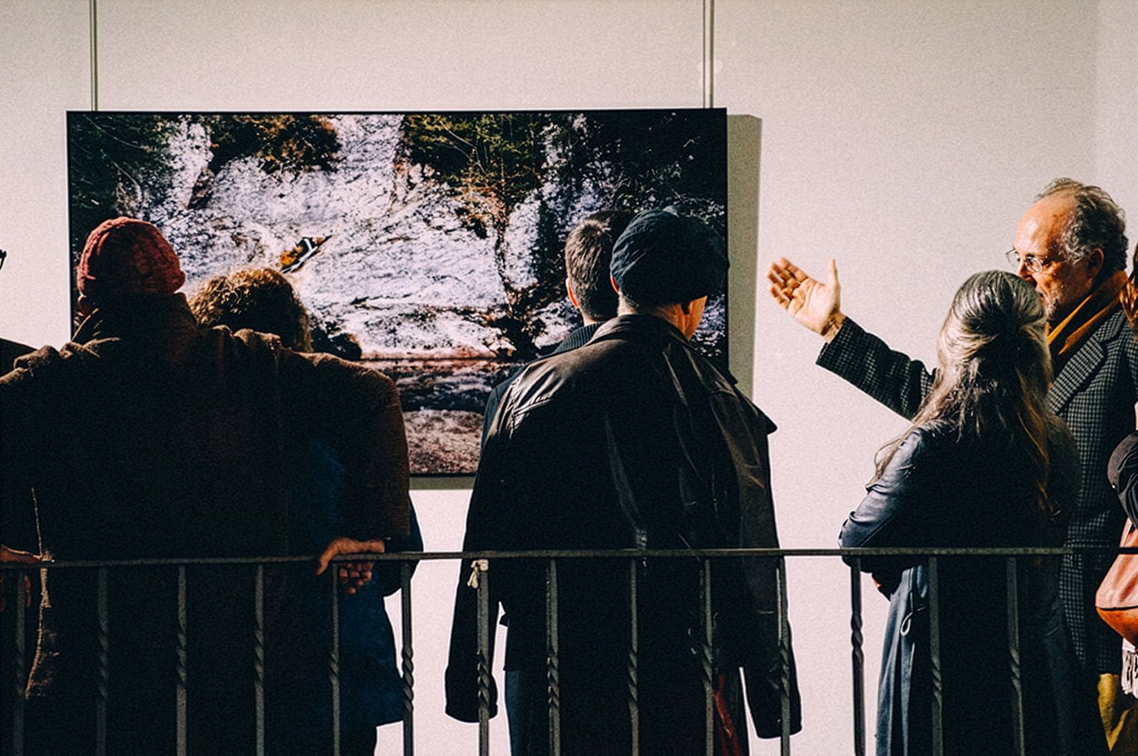 Personas de pie frente a una gran fotografía en color en una exposición, un hombre gesticulando hacia la foto.