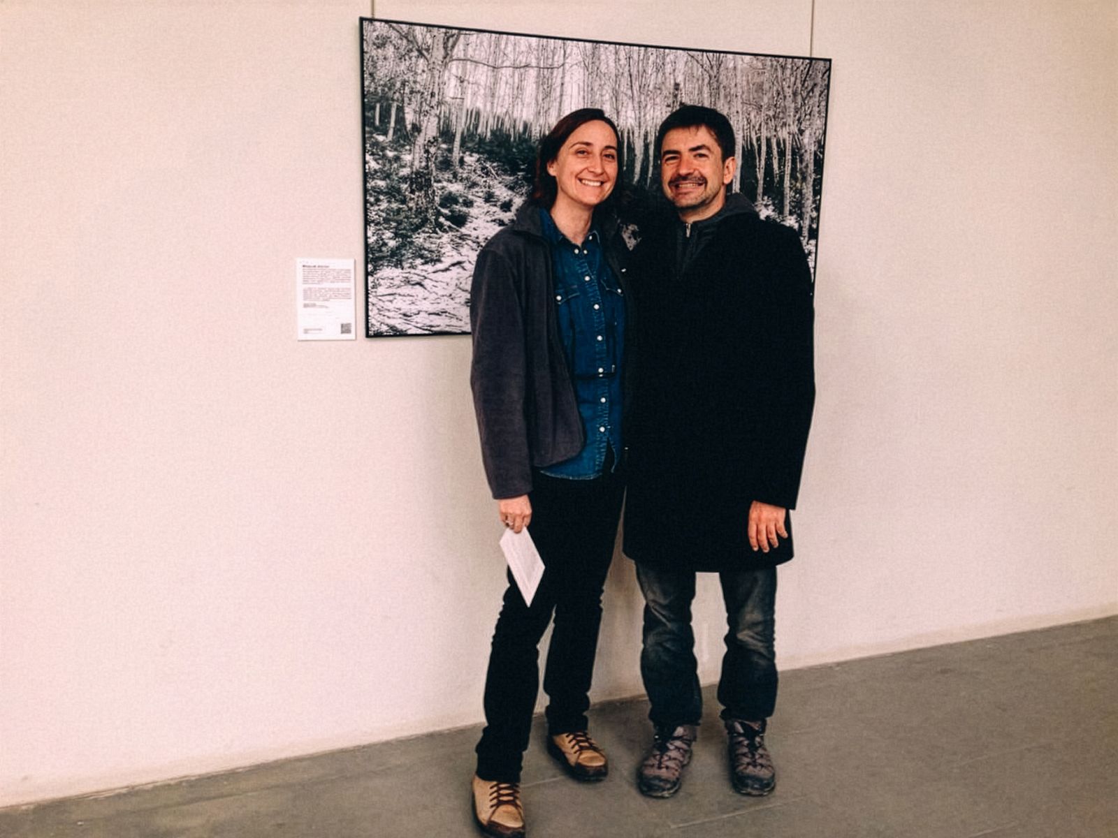 Dos personas de pie frente a una gran fotografía de un bosque nevado, sonriendo a la cámara.