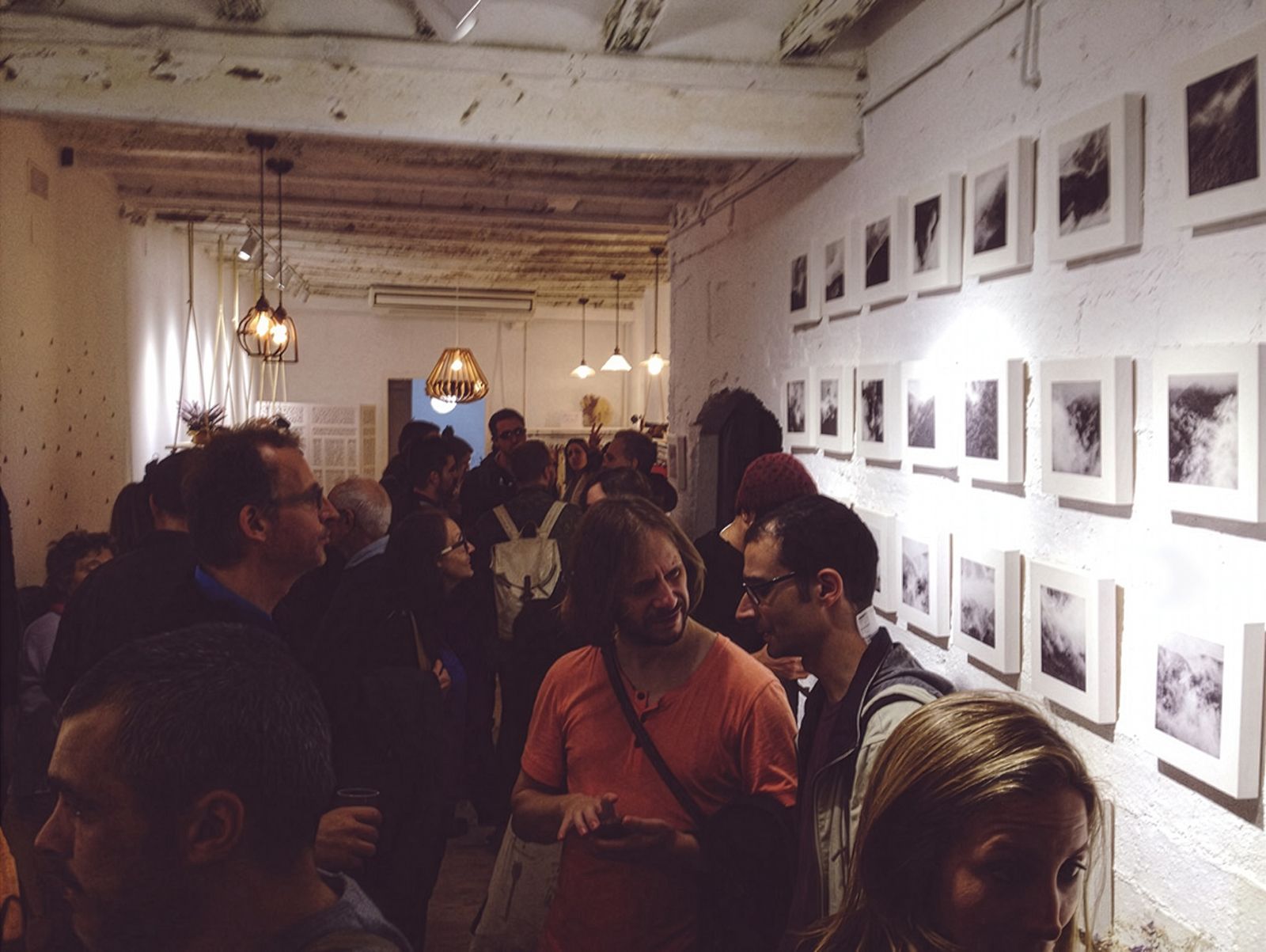 Una galeria d'art plena de gent veient fotografies en blanc i negre de núvols i boira disposades en graella en una paret de totxo blanc.